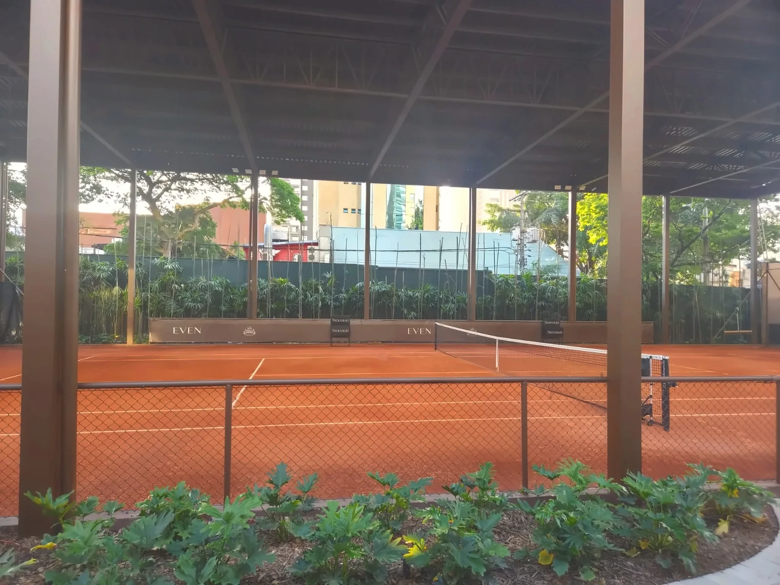 Quadra de tênis de saibro coberta, com rede e vegetação ao redor, vista da área externa à quadra.